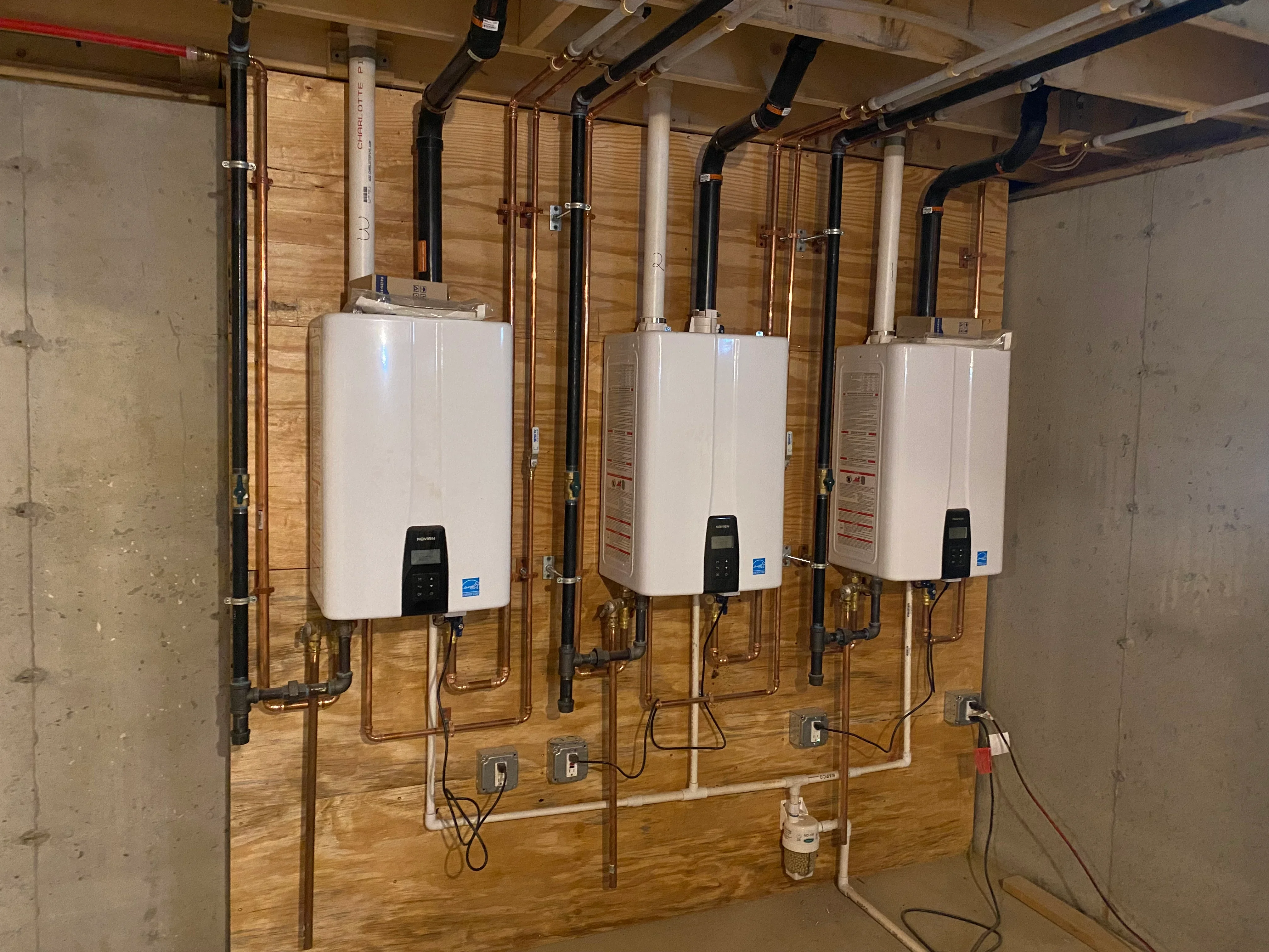 Three white tankless water heaters mounted on wall with copper and black iron piping connections
