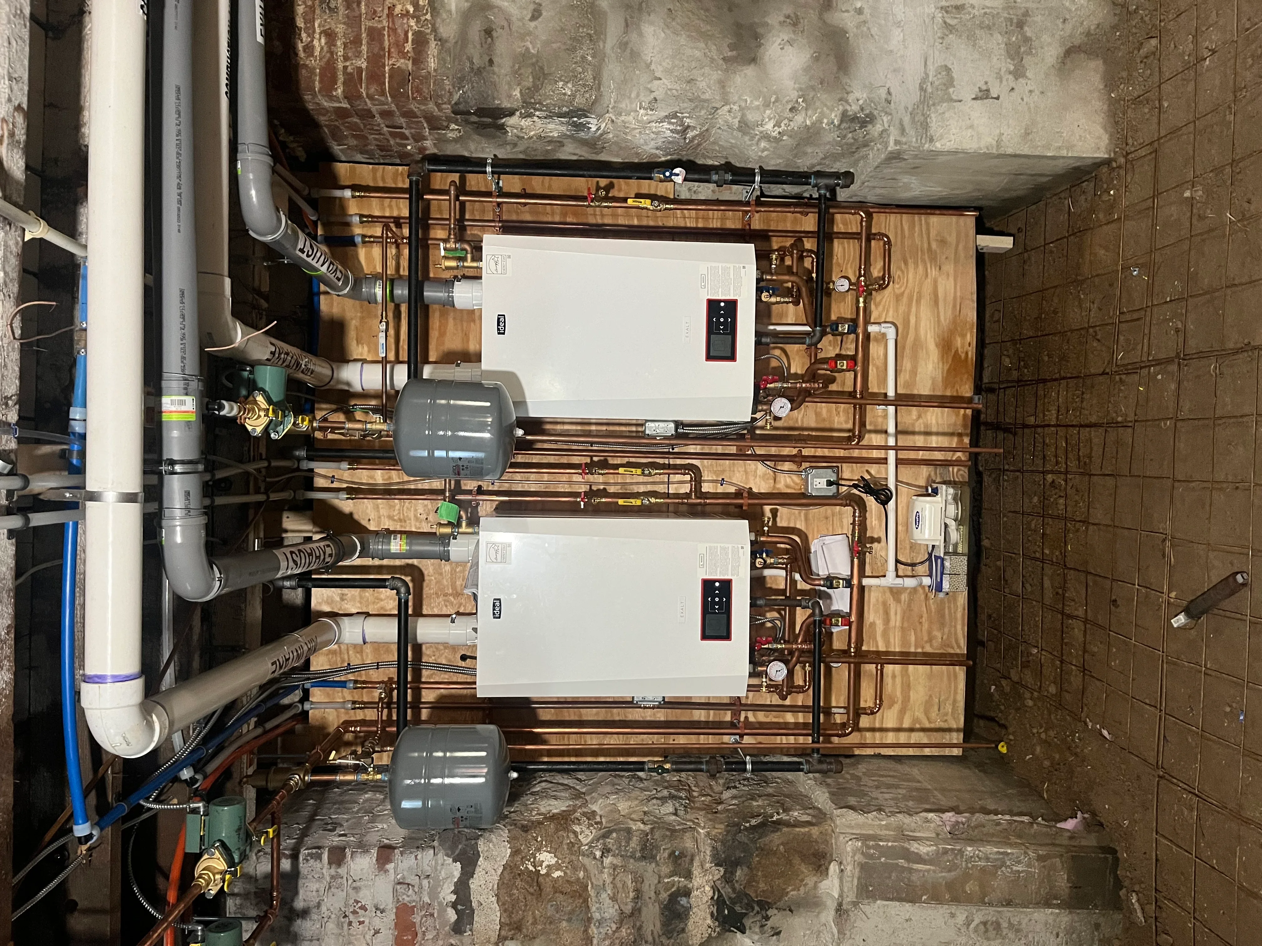 Dual tankless water heaters with copper piping, expansion tanks, and digital controls installed on wooden backing